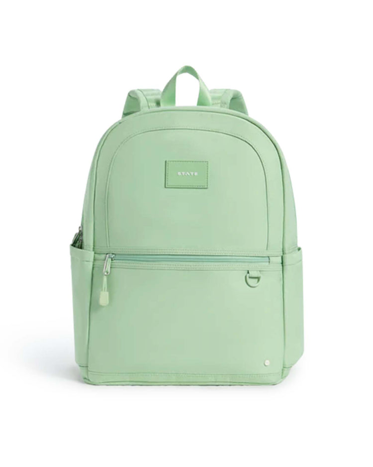 A light green Kane Double Pocket Backpack by STATE Bags, featuring a front zipper pocket, side pockets, and a top handle. The brand's label is centered on the front, and the design is simple and modern—ideal for school use.