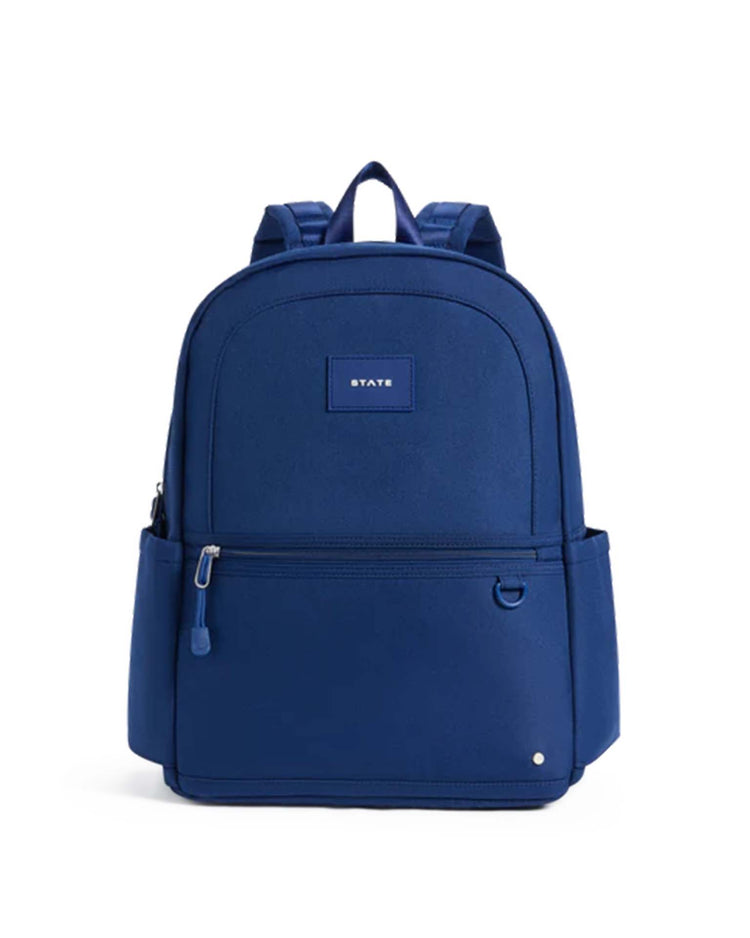 The Kane Double Pocket Backpack in Navy by STATE Bags features a simple blue design with front zippered pockets, side pockets, and a small rectangular label on the upper front displaying the word "STATE.