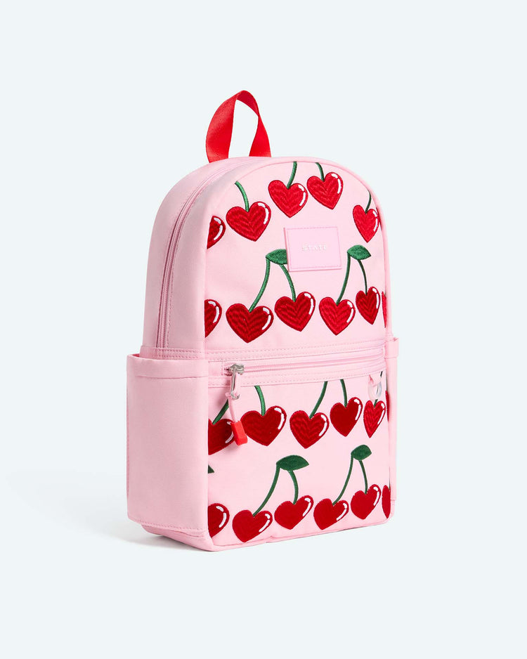 The kane mini backpack in cherry by state bags is a pink kids' backpack adorned with red heart-shaped cherry designs, featuring side pockets, a front zipper pocket, and a bold red top handle.