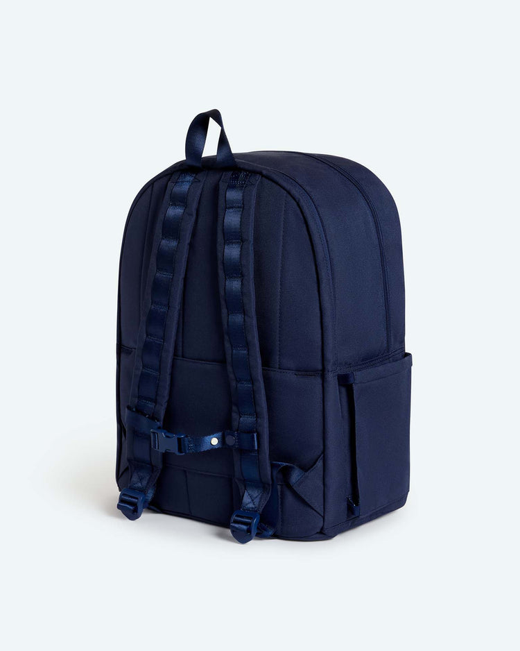 The STATE Bags Reed Backpack in midnight blue is featured from the back against a plain light background, showcasing its high-quality construction, padded shoulder straps, reinforced stitching, side pockets, and top handle.