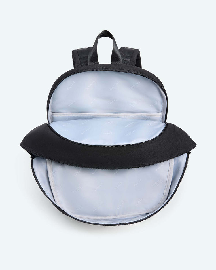 The Reed Backpack in black by STATE Bags is pictured with its main compartment unzipped, showing a light blue interior lining. This school backpack features padded shoulder straps and a top handle.