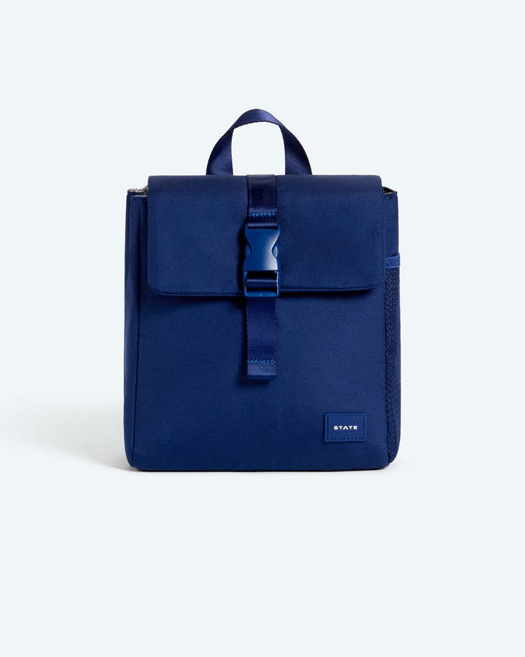 A square navy blue state bags richmond lunch sack with a top handle, front buckle closure, a side mesh pocket ideal for holding an insulated lunch sack or kids lunch box, and a small rectangular logo patch at the bottom right. The background is light gray.