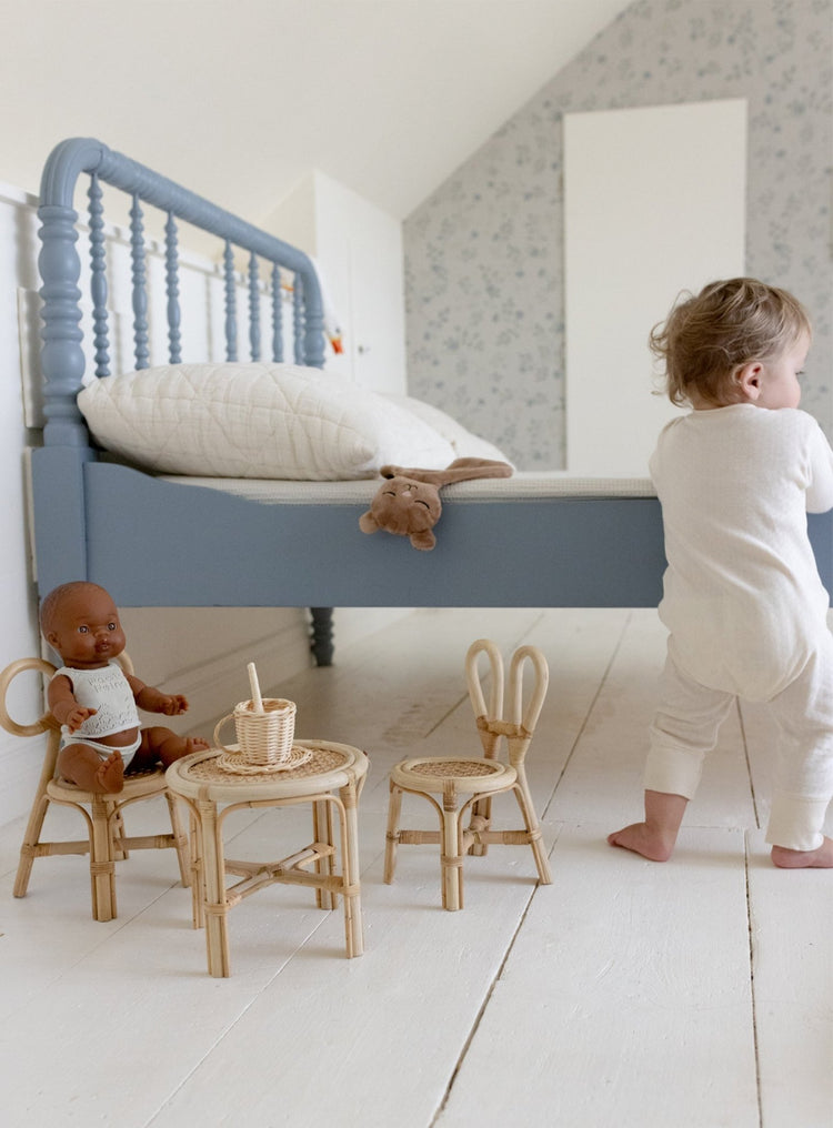 A toddler in white pajamas stands by a blue bed. On the white wooden floor, two dolls have a tea party at a small wicker table with the Poppie Mini Bunny Chair Set by poppie toys and a toy tea set.
