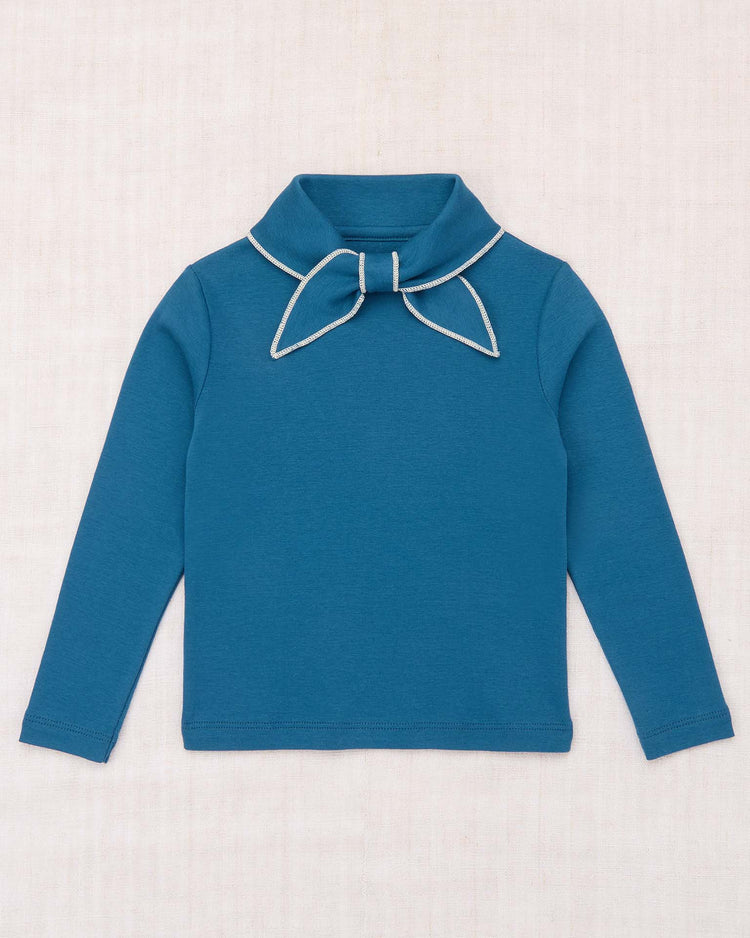 The Scout Top in Blue Smoke from misha + puff is a long-sleeve signature style crafted from organic Pima cotton, featuring a wide collar and an attached fabric bow at the neck with white trim, shown laid flat on a light, textured background.