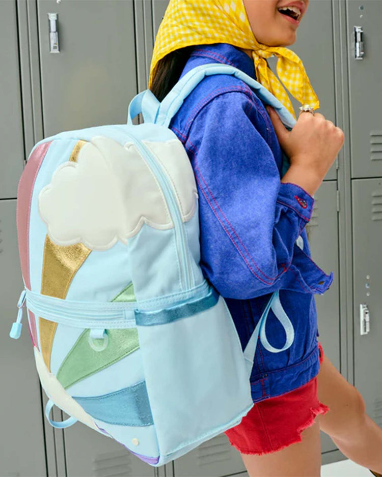 A child wearing a yellow scarf, blue jacket, and red shorts smiles while carrying the STATE Bags Kane Backpack in Rainbow Cloud in front of gray lockers.