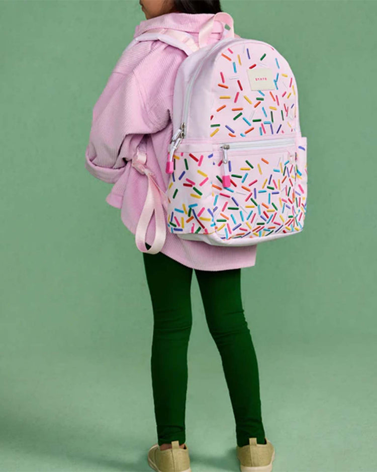 A child in a pink jacket and green leggings stands with the State Bags Kane Mini Backpack in Sprinkles on a green background.
