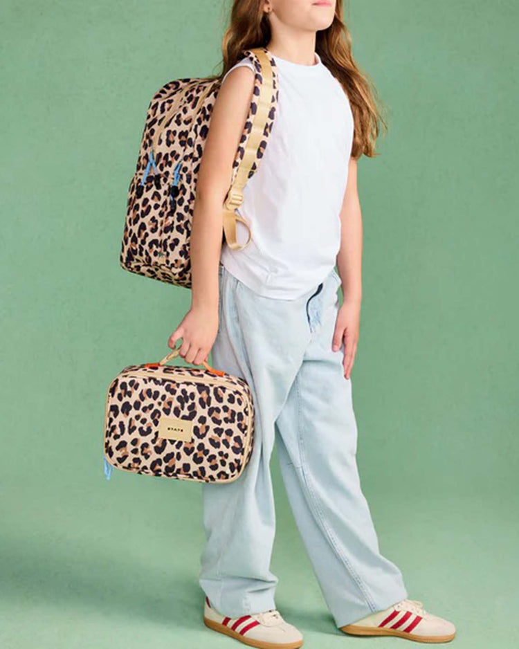 A child wearing a white sleeveless top and light blue jeans holds the STATE Bags Rodgers Lunch Box in Leopard and wears a matching backpack, standing against a green background.