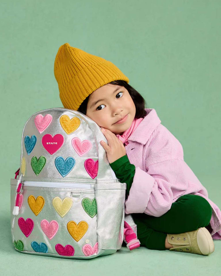 A young child in a yellow beanie and pink coat hugs the State Bags kane mini backpack in happy fuzzy hearts, sitting against a mint green background.