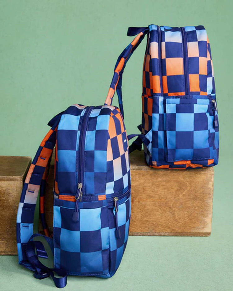 Two STATE Bags Kane backpacks in gradient checkerboard sit on wooden blocks against a green background.