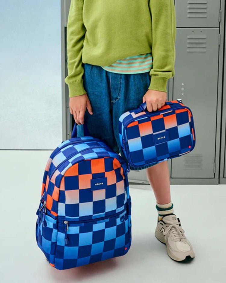 A person in a green sweater and blue shorts holds a state bags rodgers lunch box in gradient checkerboard, along with a matching backpack, while standing in front of lockers.