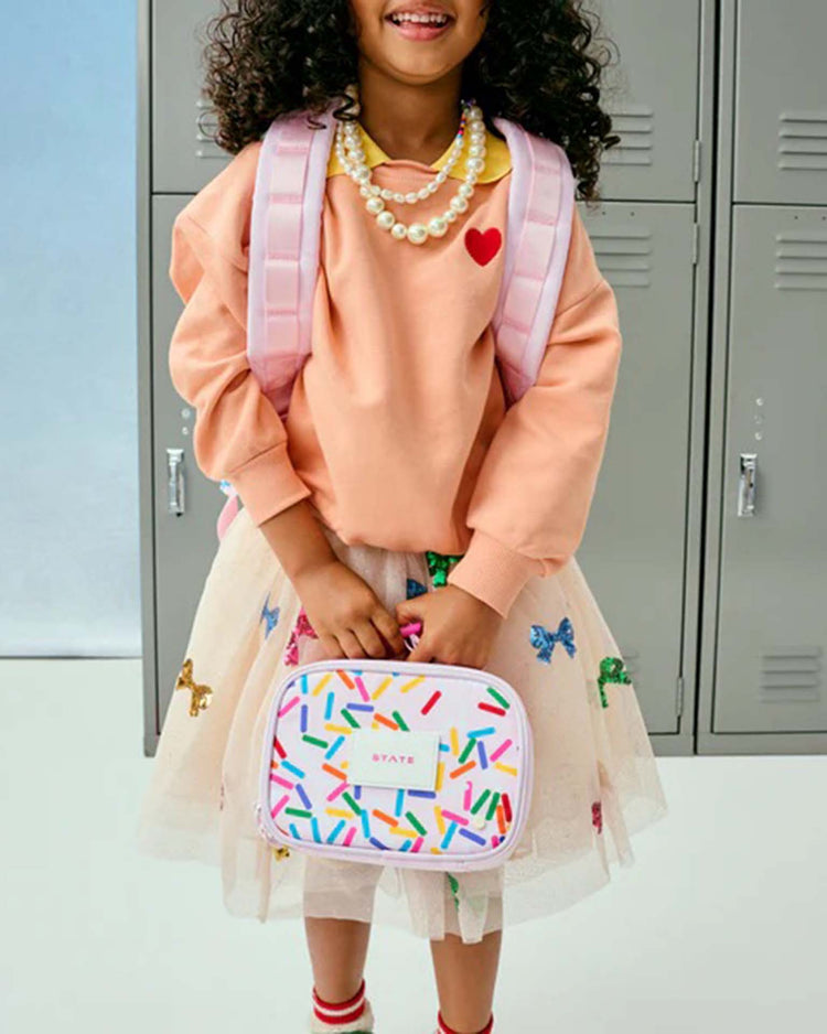 A young girl dressed in a peach sweater, pearl necklace, a colorful tulle skirt, and a pink backpack stands in front of gray lockers, holding the STATE Bags mini Rodgers Snack Pack in Sprinkles.