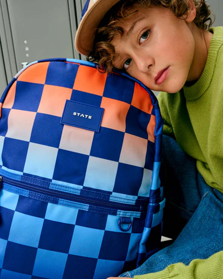 A child in a green sweater and cap leans on a STATE Bags Kane Double Pocket Backpack in Gradient Checkerboard—featuring blue, orange, and white checks, plus the STATE label—while sitting near gray lockers.