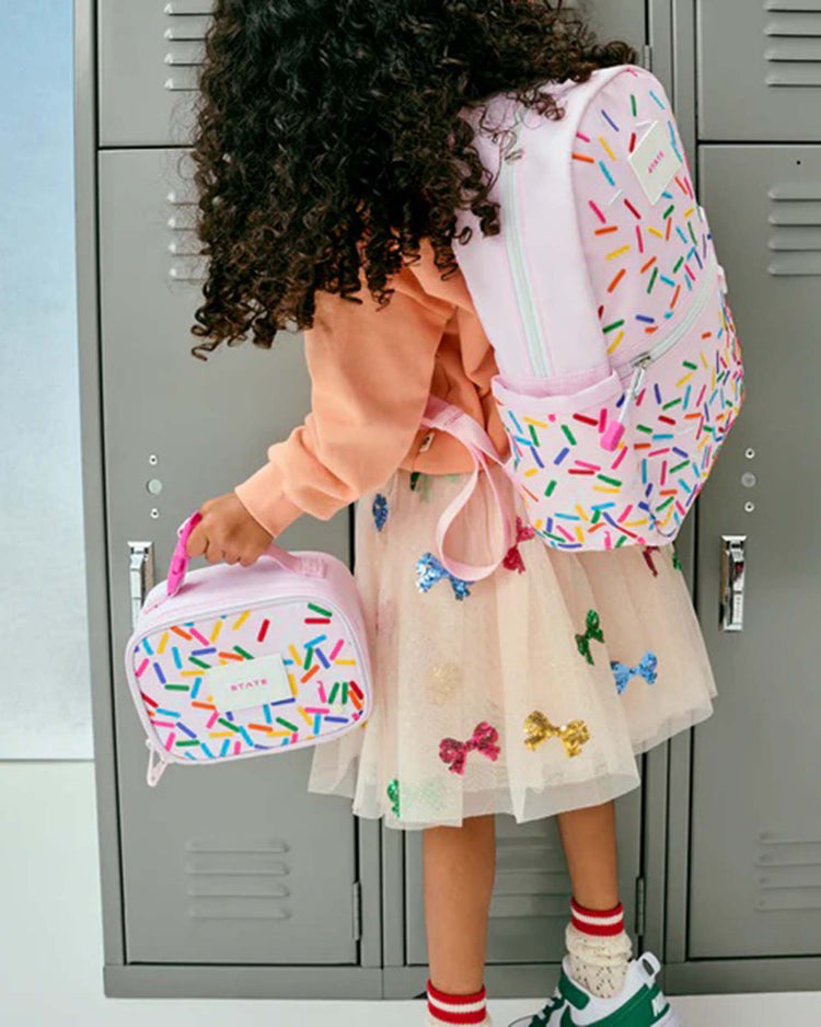 A young child with curly hair, dressed in a peach sweater and a skirt decorated with bows, stands in front of gray lockers holding the STATE Bags Mini Rodgers Snack Pack in Sprinkles along with the matching backpack.
