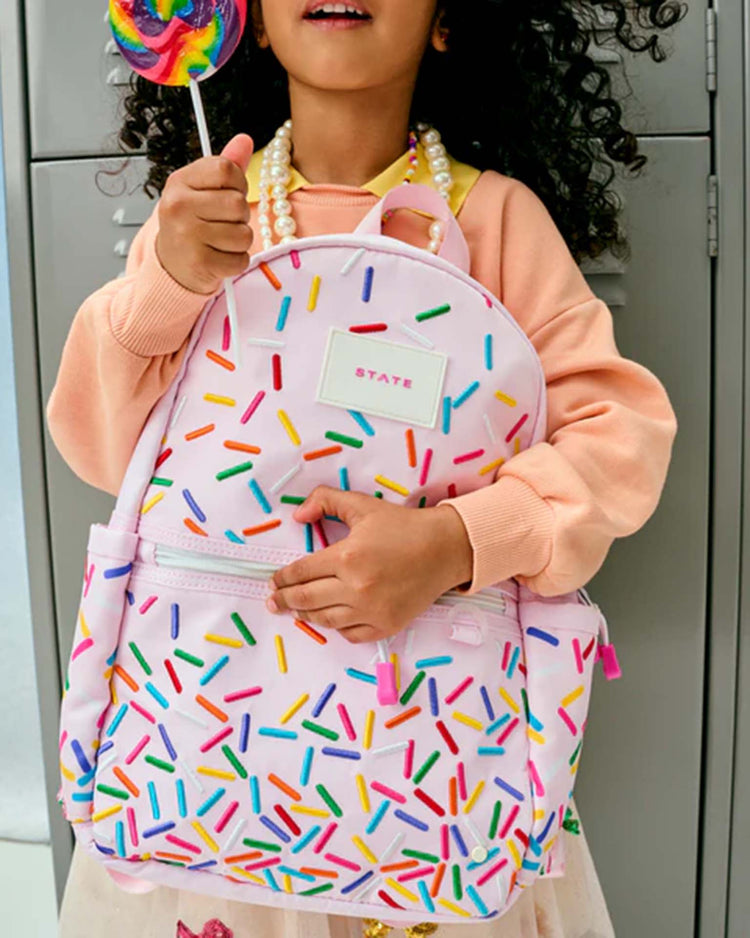 A child stands in front of lockers, wearing a peach sweatshirt and beaded necklace, holding a colorful lollipop and carrying the State Bags Kane Mini Backpack in Sprinkles.