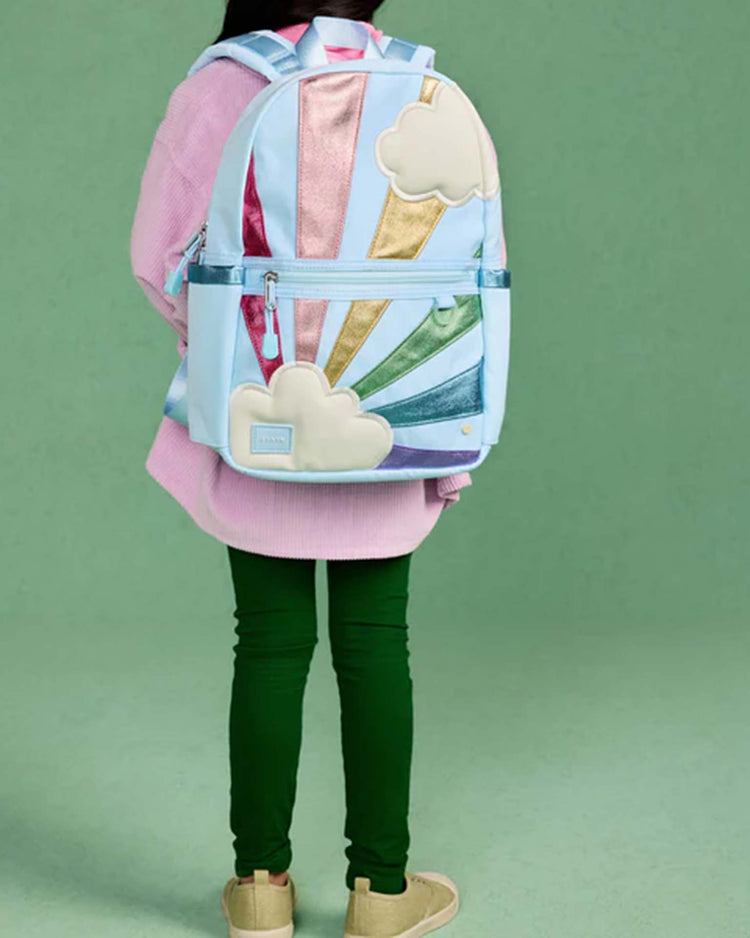 A child stands facing away, wearing the State Bags Kane Mini Backpack in Rainbow Cloud—a pastel blue kids' backpack adorned with clouds and colorful metallic rainbow stripes—against a green background.
