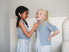 Two children dressed in blue knitwear against white bedroom backdrop. One child tying a bow on around the other's head.