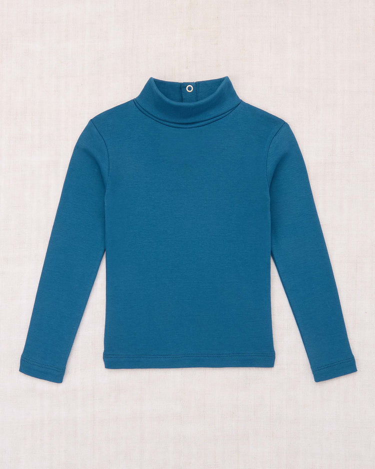 A long-sleeve turtleneck in blue smoke by misha + puff is laid flat on a light beige background. Crafted from organic Pima cotton, this women’s layering top features a minimalist design with a snap button at the back of the collar for effortless style.
