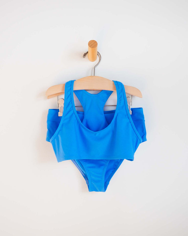 The luisa two-piece swimsuit in mare by Sunchild, featuring a sporty top and high-waisted bottoms, is neatly hung on a wooden hanger against a plain white wall.