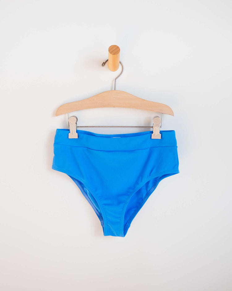 A pair of vibrant blue Sunchild "luisa two-piece swimsuit in mare" high-waisted bikini bottoms hangs on a wooden hanger against a white background, ideal for mixing and matching with your favorite swimwear styles.