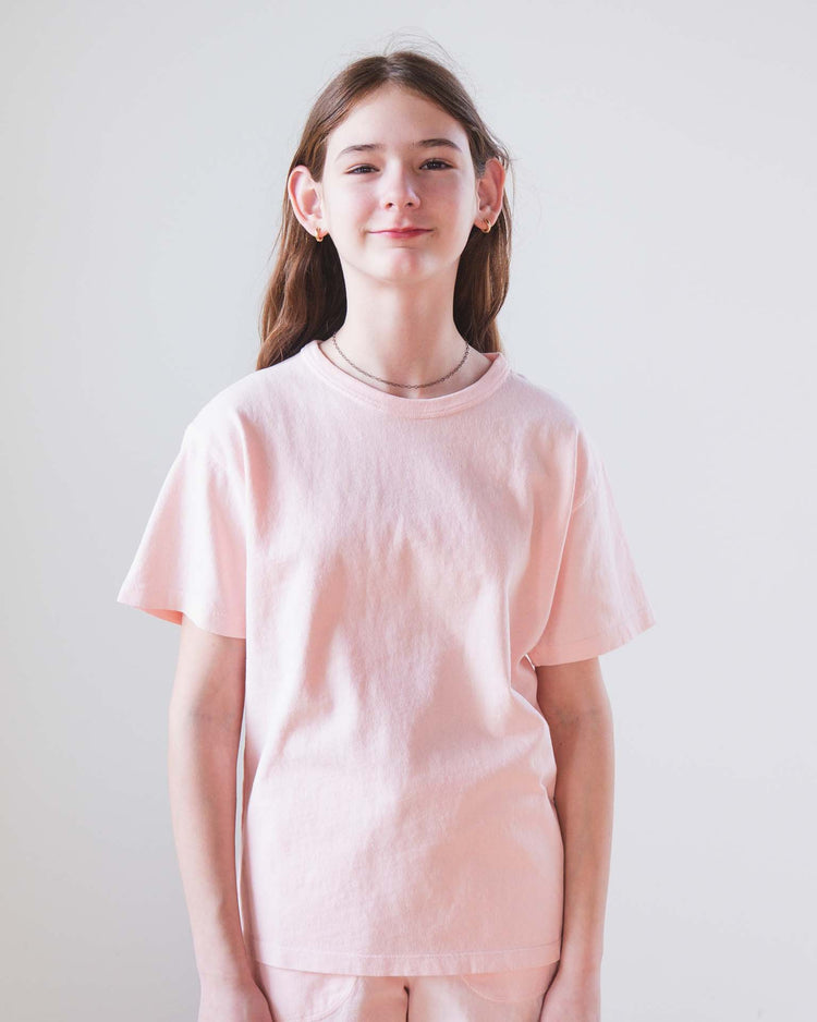 A young person with long brown hair stands against a plain light background, wearing the sunchild kazan tee in baby and looking at the camera with a slight smile.