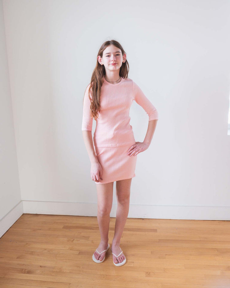 A girl with long brown hair stands indoors on a wooden floor against a white wall, wearing the Sunchild Paz skirt in baby with matching sandals. She has a relaxed expression.