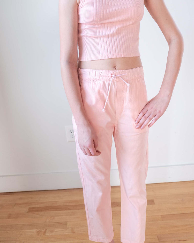 A person stands indoors on a wooden floor, wearing sunchild's mustang pants in baby along with a light pink sleeveless ribbed crop top, one hand resting on their hip.