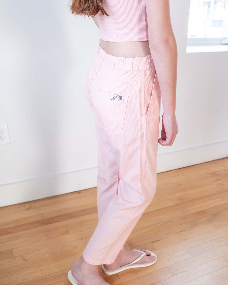 A person stands indoors on a wooden floor, wearing sunchild's mustang pants in baby—100% cotton with an elastic waistband and back pocket patch—paired with a pink sleeveless top and white flip-flops.