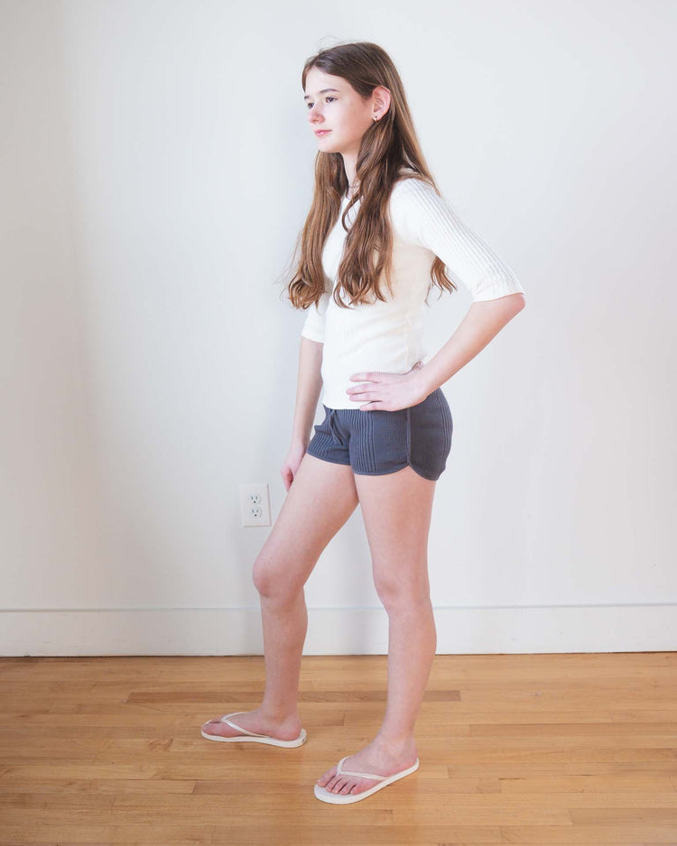 A young woman with long brown hair stands sideways indoors on a wooden floor, wearing a white top and sunchild rio shorts in caviar with a drawstring waist, plus white flip-flops, hands on hips against a plain white wall.
