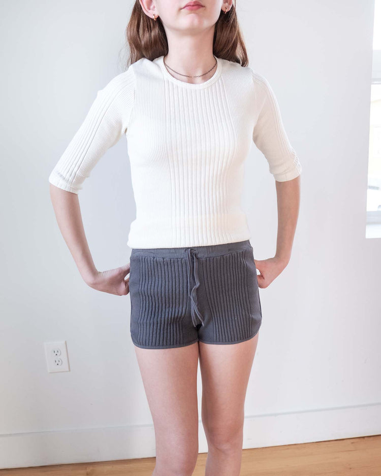 A person stands indoors wearing a white ribbed 3/4 sleeve top and sunchild rio shorts in caviar with a drawstring waist, hands on hips. The background features a white wall, window, and power outlet.