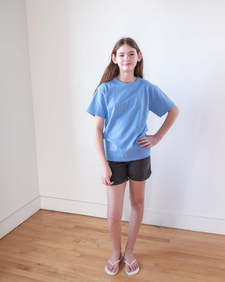 A young Sunchild with long brown hair stands indoors on a wooden floor, wearing a blue T-shirt, Sunchild hunter shorts in caviar with a drawstring waistband, and white sandals. One hand is on her hip as she poses against a plain white wall.