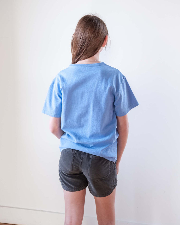 A person with long brown hair, wearing a light blue t-shirt and Sunchild hunter shorts in caviar featuring a drawstring waistband, stands facing away from the camera against a plain white wall.