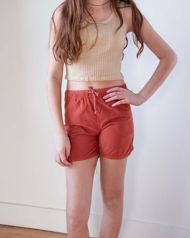 A young person with long brown hair wears a sunchild cruz tank in craie and rust-colored shorts, standing indoors with one hand on their hip against a plain white wall.