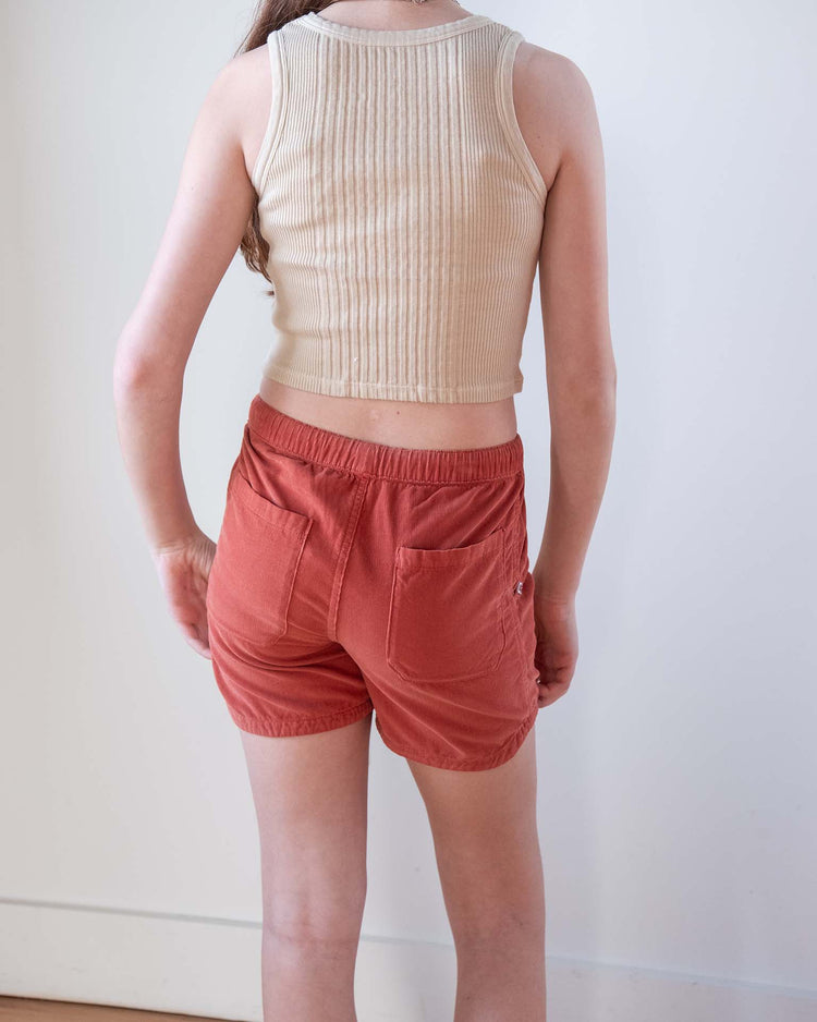 A person indoors faces away, wearing the sunchild cruz tank in craie and rust shorts with back pockets. The setting is a plain white wall and light wood floor—a relaxed look from sunchild.