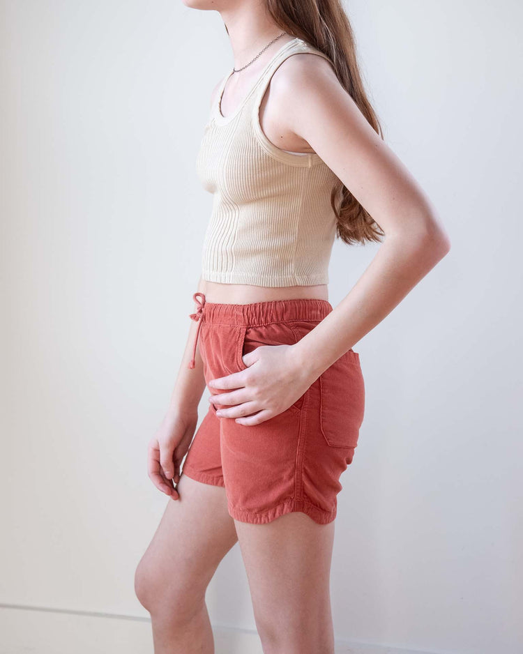 A person with long brown hair wears the sunchild cruz tank in craie and high-waisted, rust-colored drawstring shorts, standing sideways against a plain light background.