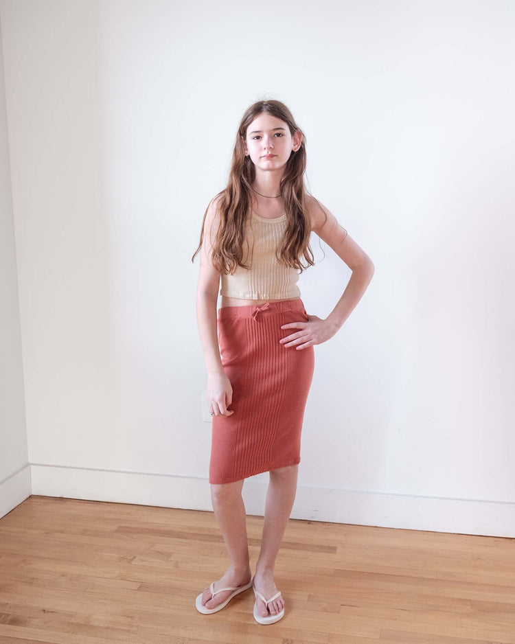 A young woman with long brown hair stands indoors on a wooden floor, wearing a beige sleeveless top, the Sunchild siuna skirt in cacao (100% cotton), and white flip-flops, against a plain white wall—a true Sunchild look.