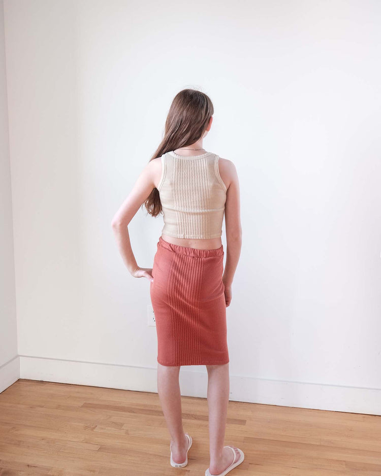 A woman with long brown hair stands indoors facing a white wall, wearing the Sunchild cruz tank in craie, a high-waisted rust-colored skirt, and white sandals on a light wooden floor.