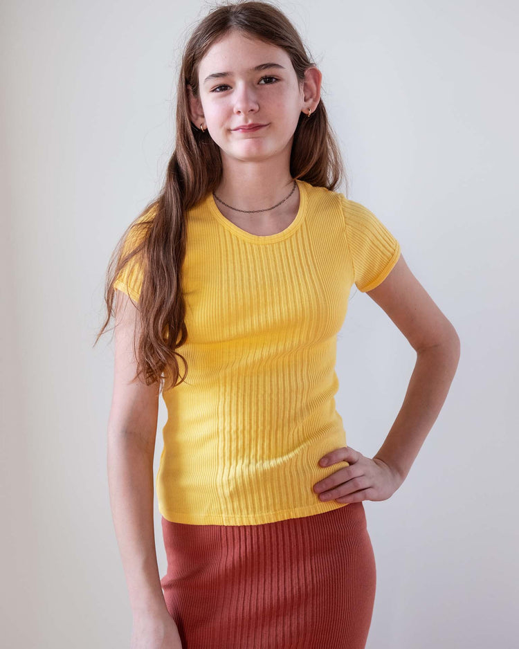A young person with long brown hair stands against a plain light background, wearing a fitted short-sleeve sunchild loutro tee in napoli and a rust-colored ribbed skirt, one hand on their hip and a slight smile.