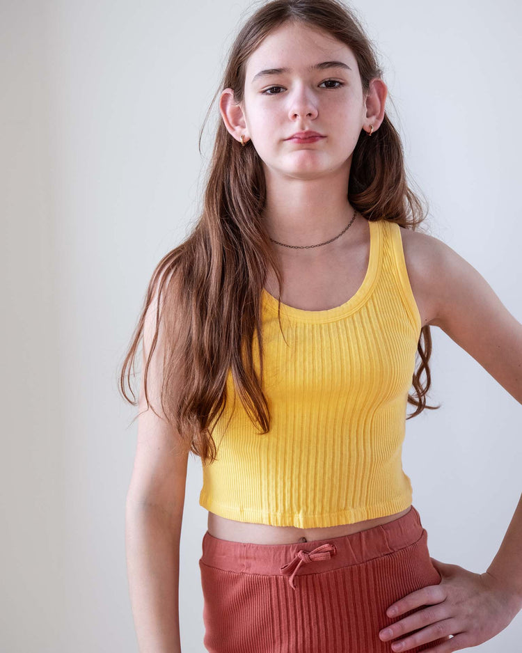 A young person with long brown hair wears the sunchild cruz tank in napoli and rust-colored pants, standing against a plain light background with one arm resting on their hip.