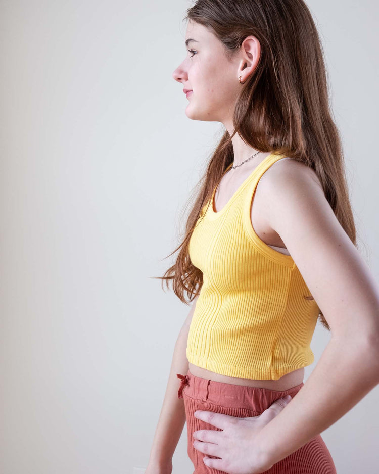 A young woman with long brown hair stands in profile against a plain light background, wearing the sunchild cruz tank in napoli and high-waisted rust-colored pants, with her hand resting on her hip.