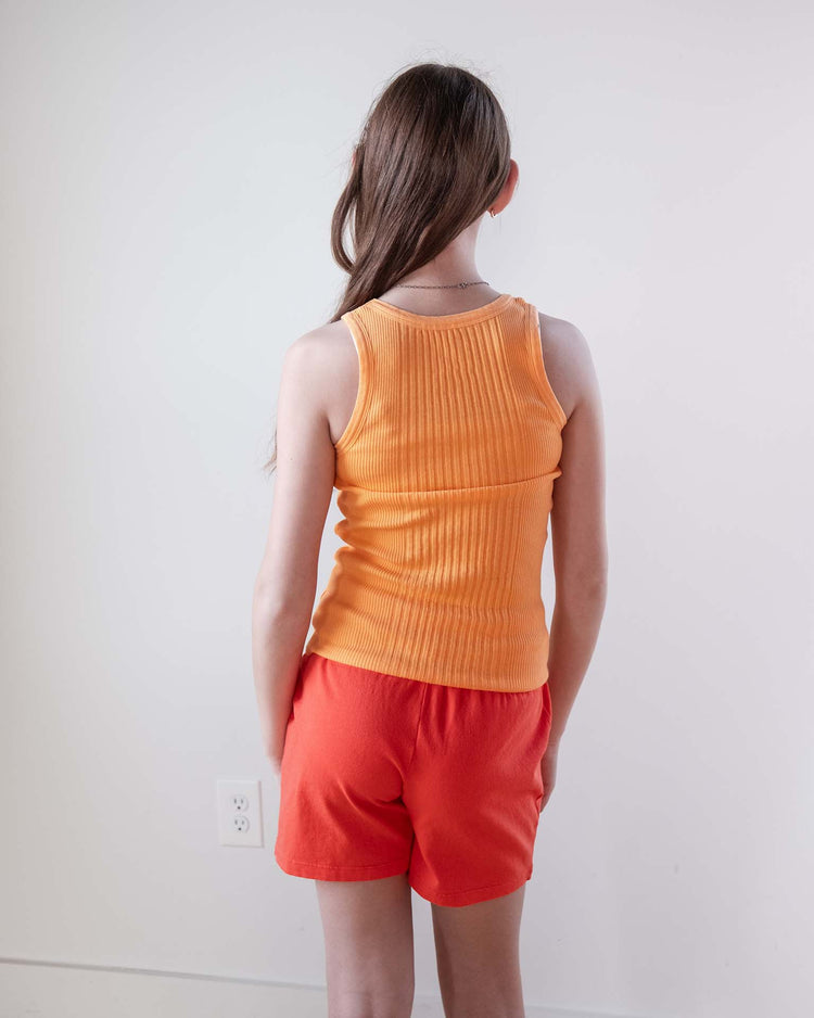 A young girl with long brown hair faces away from the camera, wearing an orange ribbed sleeveless top and Sunchild merida shorts in tangerine with a drawstring waist, set against a plain white wall.