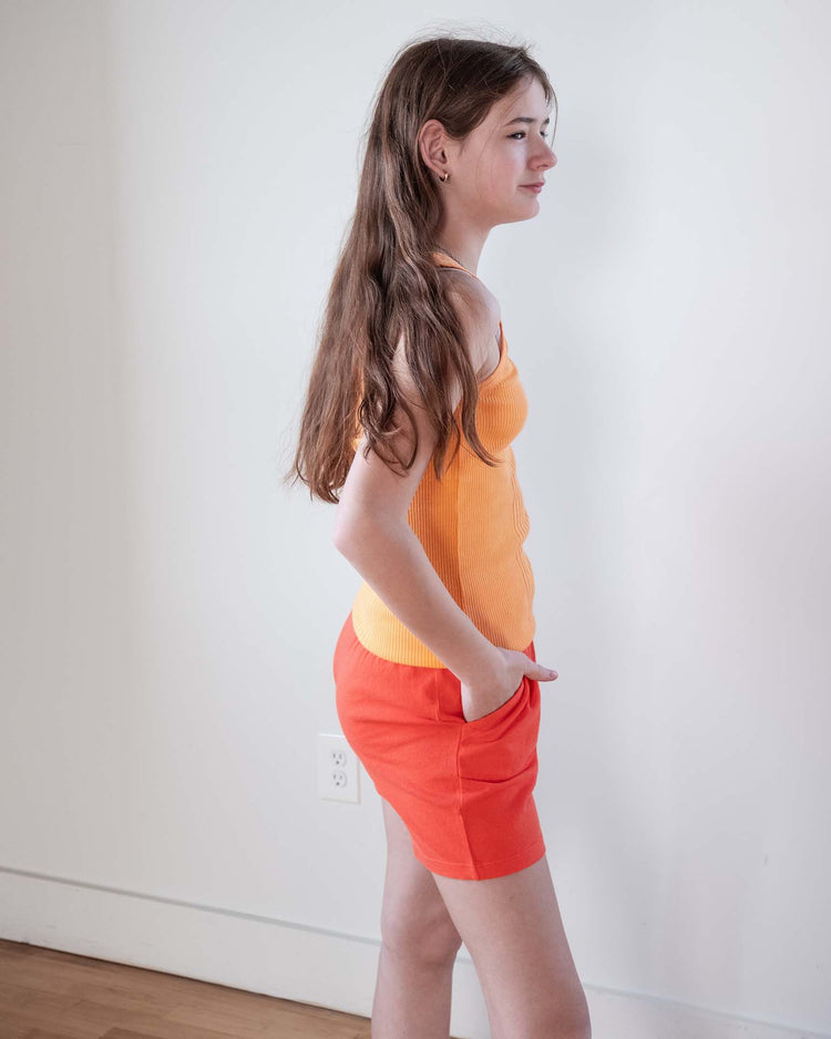 A young person with long brown hair stands sideways against a plain white wall and wooden floor, wearing the Sunchild fernando tank in inca and red-orange shorts with hands in pockets.