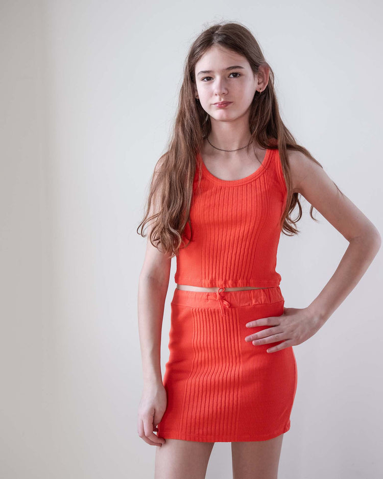 A young woman with long brown hair poses against a plain background, wearing a sleeveless ribbed crop top and the matching Sunchild paz skirt in tangerine—an eye-catching summer look. One hand rests on her hip as she stands neutrally.