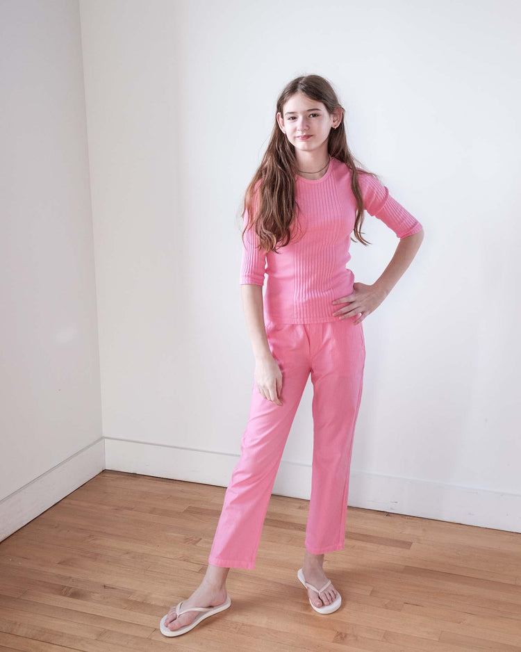 A young person with long brown hair in a pink ribbed top and sunchild's mustang pants in glamour with a drawstring waist stands on wooden floors by white walls, completing the look with classic white flip-flops.