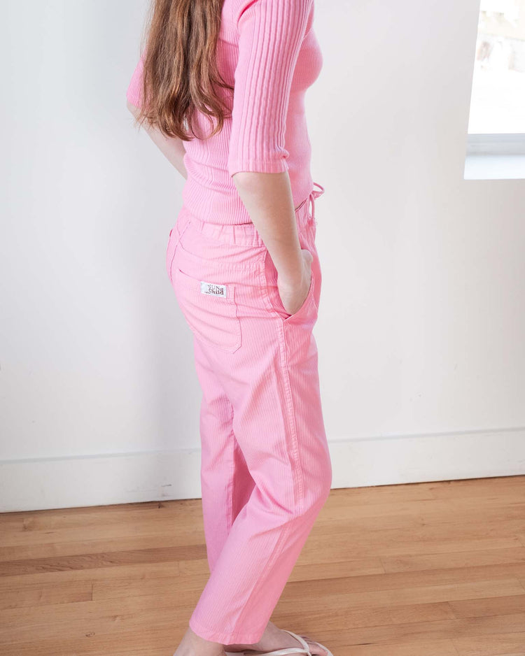 A person with long brown hair, wearing a pink ribbed top and sunchild mustang pants in glamour with a drawstring waist and hands in pockets, stands on a wooden floor near a white wall and window.