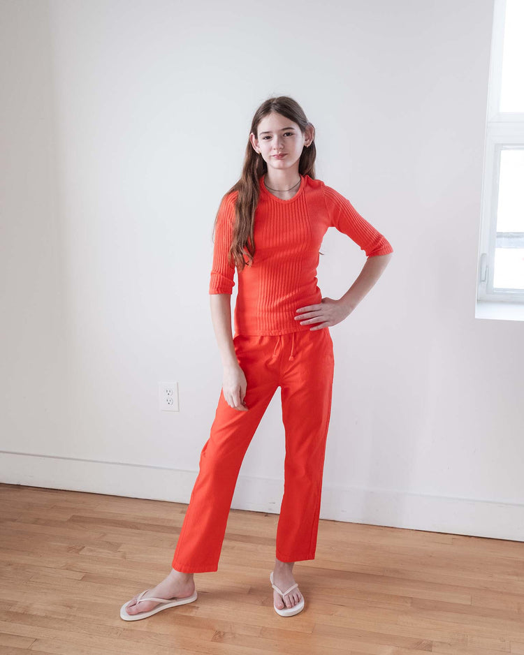 A young woman with long brown hair stands indoors on a wooden floor, wearing the Sunchild puebla tee in tangerine and matching pants, inspired by Marseille French style. She poses with one hand on her hip and a neutral expression.