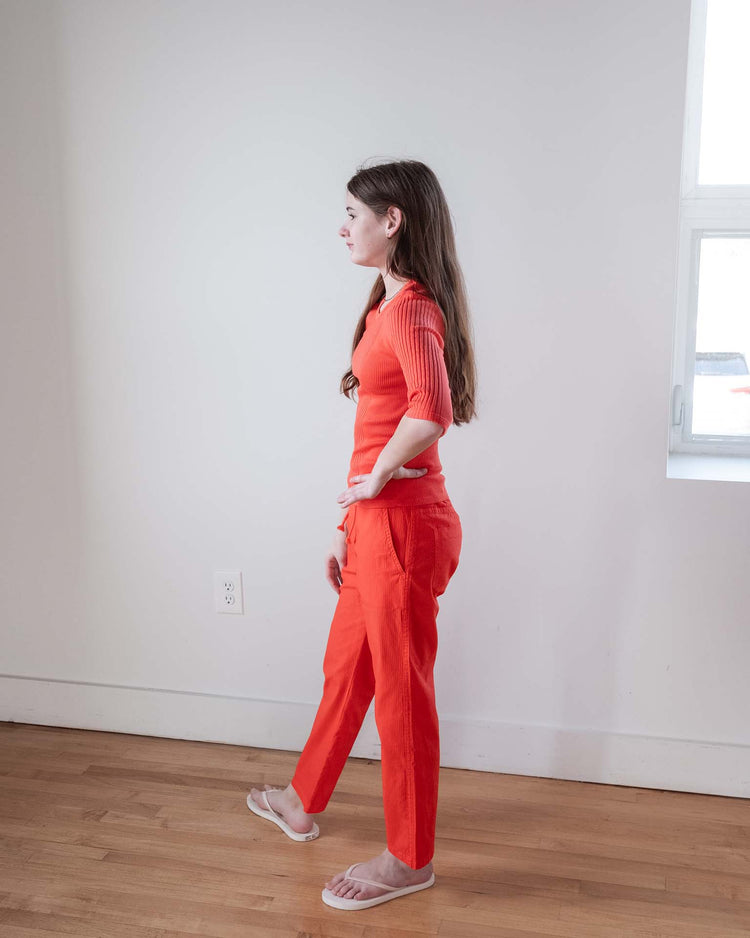 A woman with long brown hair stands in profile indoors, wearing the Sunchild Puebla tee in tangerine, matching orange pants, and clear sandals. She poses against a white wall with a window on her right.