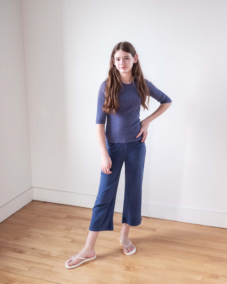 A young girl with long brown hair stands on a wooden floor indoors, posing confidently in the sunchild puebla tee in obscur, blue cropped pants, and white sandals against a white wall.