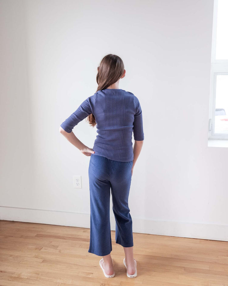 A woman with long brown hair stands indoors facing a white wall, wearing the sunchild puebla tee in obscur and matching blue pants. One hand is on her hip, white shoes complete her look, and a small window appears on the right.