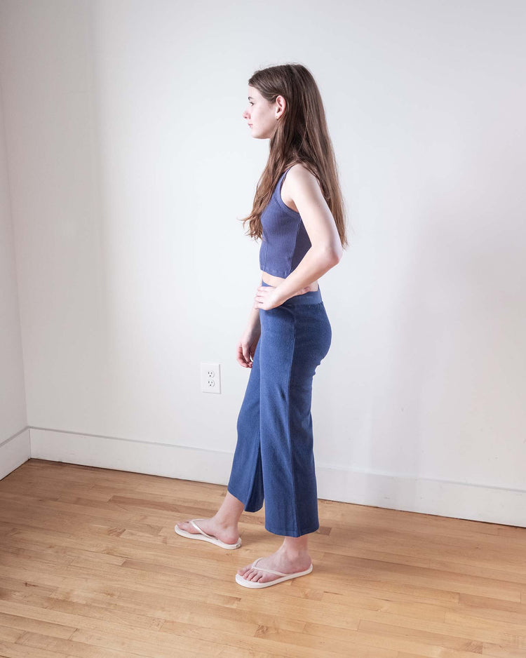 A young woman with long brown hair stands sideways in a room with light walls and wooden floors, wearing the sunchild cruz tank in obscur, blue pants, and flip-flops. Her hand rests on her hip as she looks ahead.