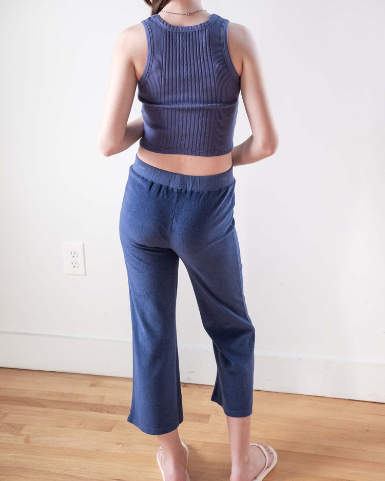 Someone stands indoors on a wooden floor, facing away, wearing a ribbed navy tank top, Sunchild’s bluff pants in obscur, and white flip-flops.